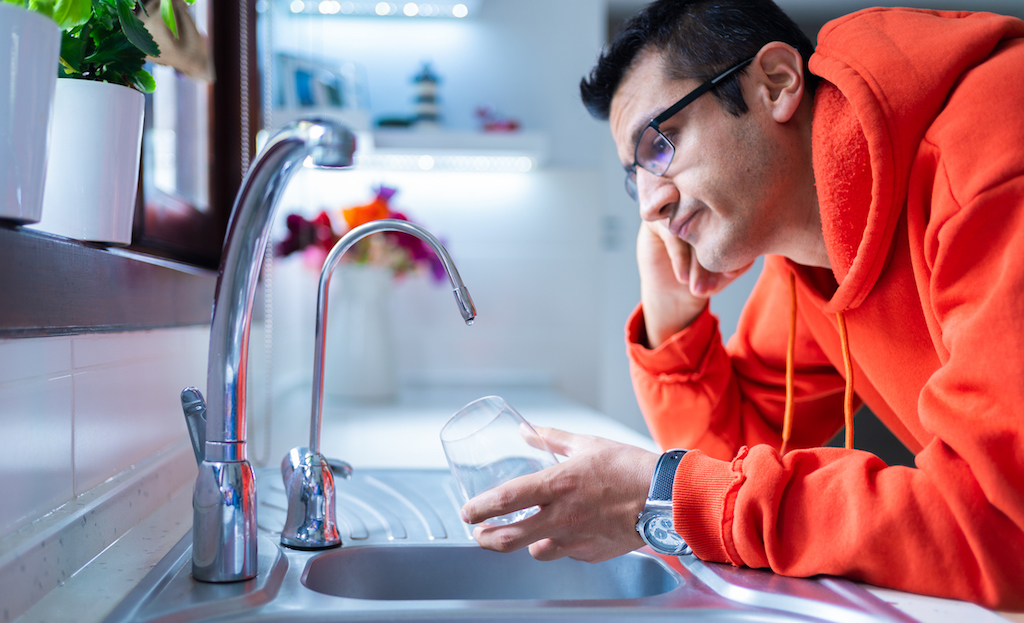 Man in orange sweatshirt holding a glass under faucet, no water due to plumber frozen pipes. 
